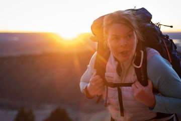 Erin Parisi is looking at the camera with the sun behind her and a backpack on her back. There's a mountain range in the distance.