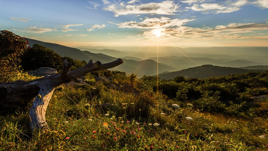 A photograph taken from a high-elevation vantage point, looking out at the sun over a mountain range. 
