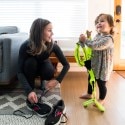 A woman with brown hair kneels down to lace her running shoes, while smiling at her toddler-aged daughter, who is next to her, holding a reflective running vest and also smiling.