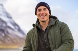 Chris Burkard is wearing a beanie and a green coat and is smiling at the camera. A mountain range is behind him, out of focus.