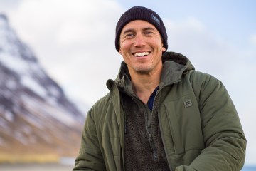 Chris Burkard is wearing a beanie and a green coat and is smiling at the camera. A mountain range is behind him, out of focus.