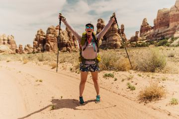 Miranda is holding hiking poles with her arms outstretched smiling into the camera. There are rock formations behind her.