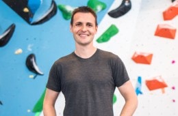 David Sacher stands in front of an indoor rock wall smiling at the camera with his hands on his hips.