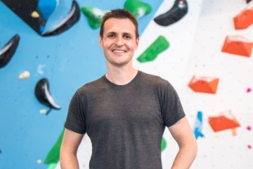 David Sacher stands in front of an indoor rock wall smiling at the camera with his hands on his hips.