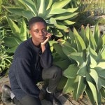 A woman in a navy sweater and black jeans kneels in front of a group large aloe plants.
