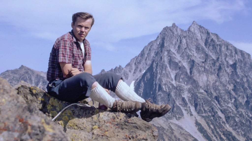 Dave Chantler on Ingalls Peak with blue skies and mountain peaks in the background. He is wearing a short sleave red plaid shirt, blue grey knickers, tall white socks and brown leather mountaineering boots.