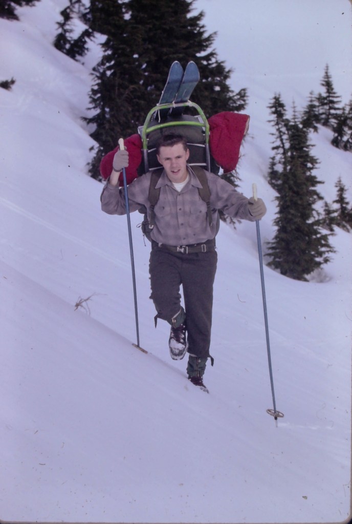 Dave Chantler is climbing up a snowy slope with pine trees in the background. He is using ski poles and carrying a large metal frame pack with skis. He is wearing brown gloves, brown pants with belt and a long sleeve brown plaid shirt.