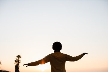 A woman wearing an orange REI Co-op Flash Jacket has her arms out and is silhouetted by the setting sun.
