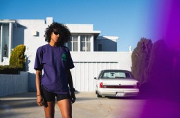 A woman wearing sunglasses, a purple shirt and black compression running shorts stands in front of a white, modern house with a classic white car in the driveway.