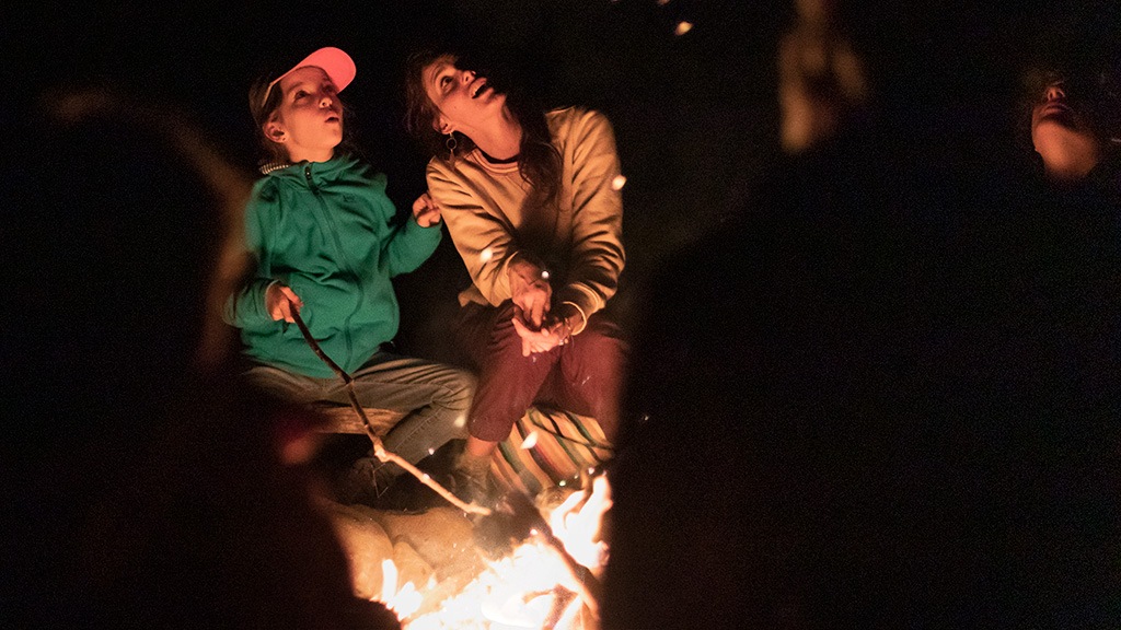 Two people gaze up in the light of a campfire