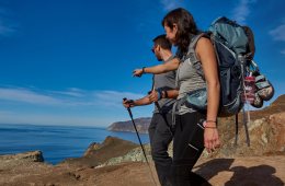 Two people hiking along a cliff look out to blue waters