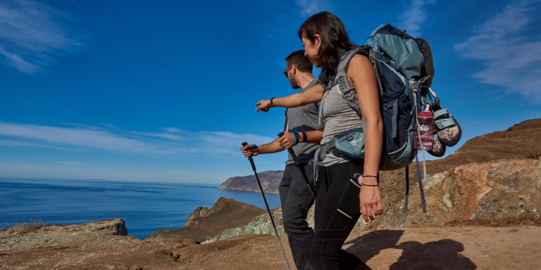 Two people hiking along a cliff look out to blue waters
