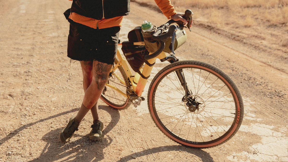 A rider stands next to a gravel bike on a dusty, dirt road.