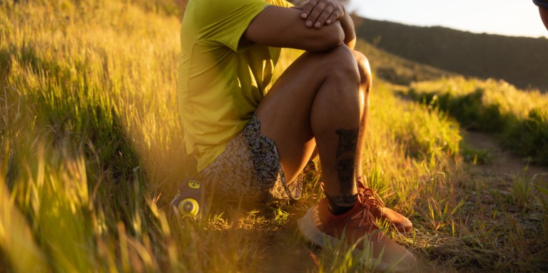 A person sitting on a grassy hill in a pair of red REI Co-op Swiftland MT Trail-Running Shoes.