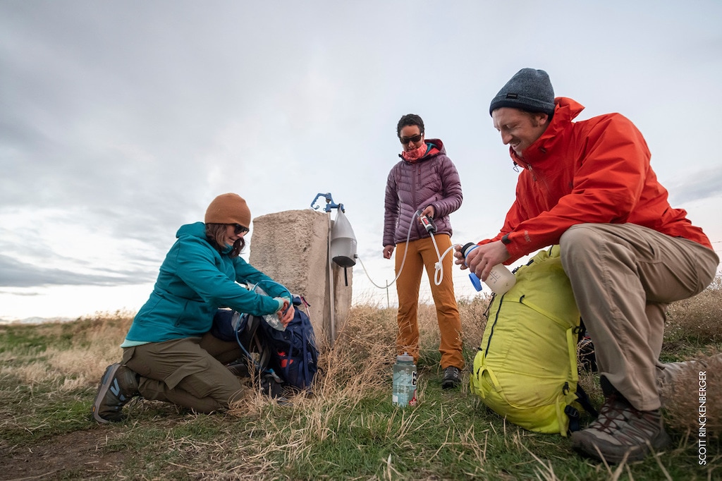 Three people in hiking gear filling up a water bottle from a pump, using their MSR Guardian Gravity Purifier.