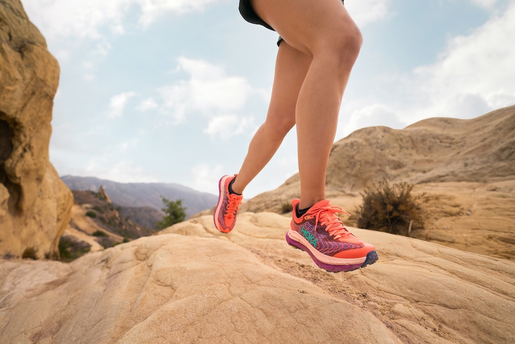 A person running across a rocky desert landscape in the HOKA Mafate Speed 4 trail running shoes in a pink color. 