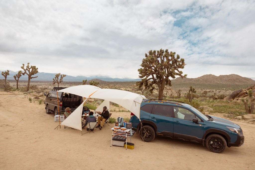 A group of car campers sit under the shade of a tarp shelter.