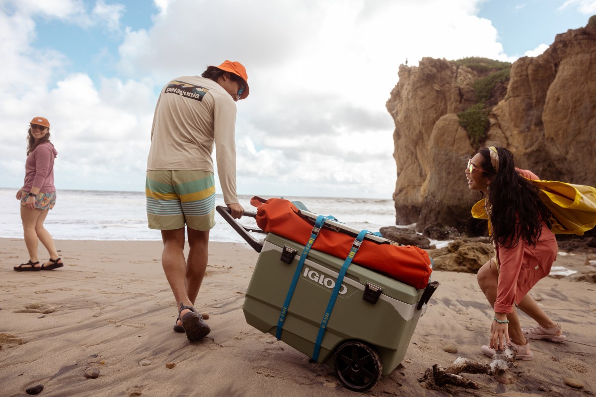 Several young people walk along a sandy beach. One of them is pulling a large cooler on wheels.