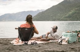 Two people sit on a picnic blanket on a beach. A cooler is next to them.