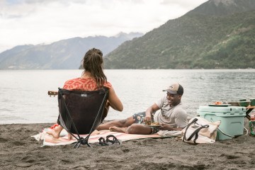 Two people sit on a picnic blanket on a beach. A cooler is next to them.