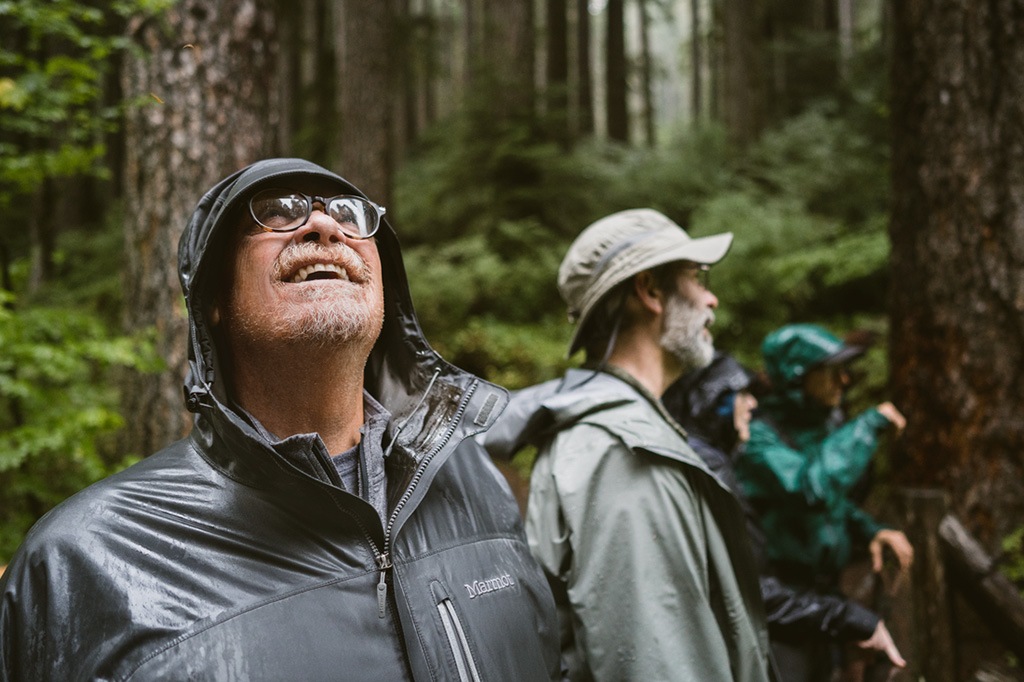 Two people wearing rain gear in a forest, one is looking up at the sky