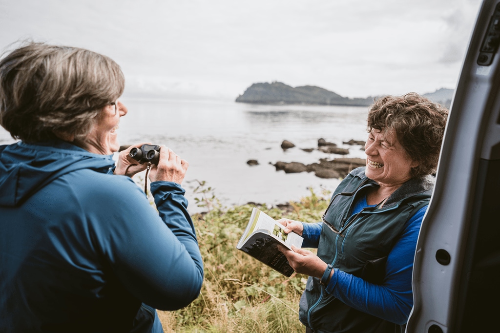 Two people laughing on a hike; one is holding binoculars and the other is holding a guidebook.