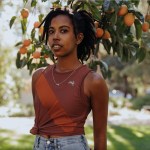 Portrait of Uncommon Path contributor Kamilah Journét wearing a burgundy singlet and jeans, standing in front of an orange tree.
