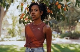 Portrait of Uncommon Path contributor Kamilah Journét wearing a burgundy singlet and jeans, standing in front of an orange tree.