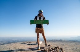 Alan Ortega in running gear on the top of a mountain holding a sign that says "Potato Mountain." There are also several potatoes at his feet.