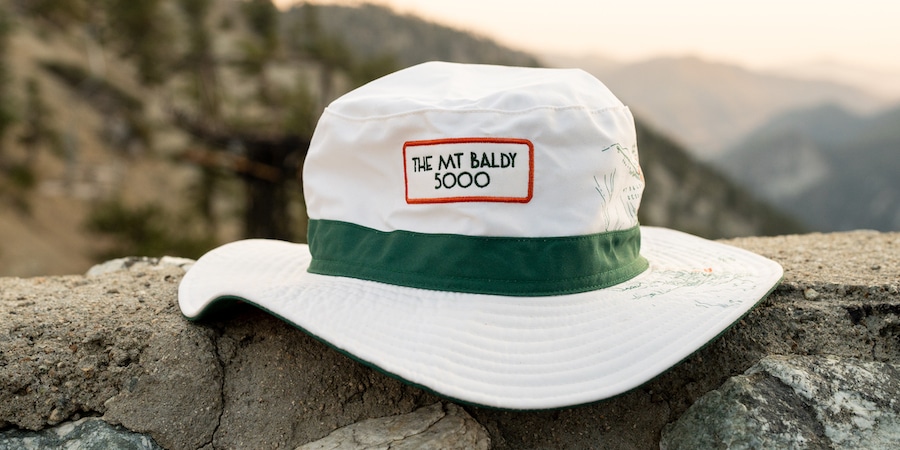 Perched on a mountain ridge, a white bucket hat with a green band around around the base of the cap and the words 