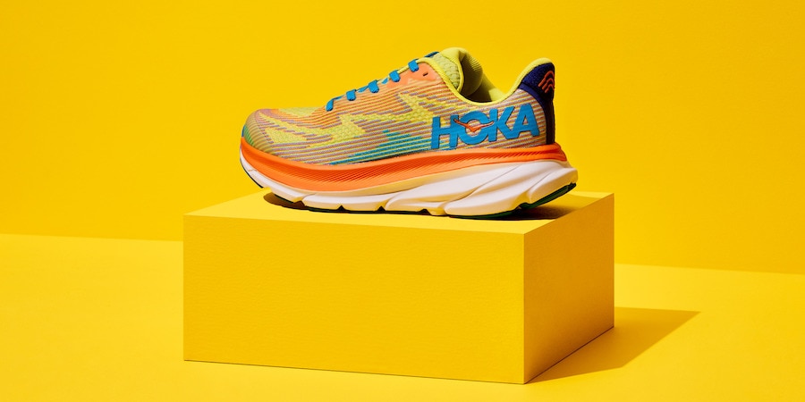 A single, multi-colored HOKA Clifton 9 Road-Running Shoe on a yellow platform against a yellow backdrop.