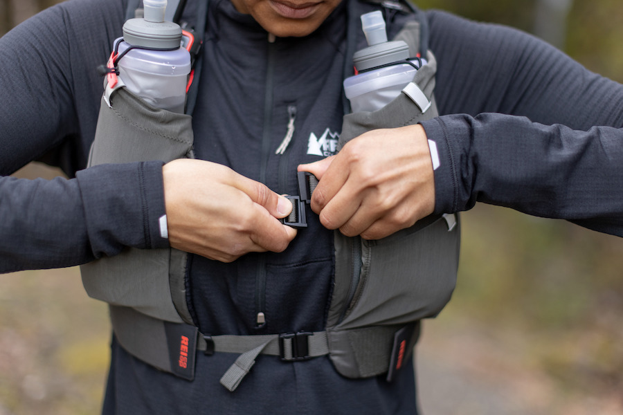 A person buckling the top strap of their REI Co-op Swiftland TT Hydration Vest, which has two bottles in the chest pockets. 