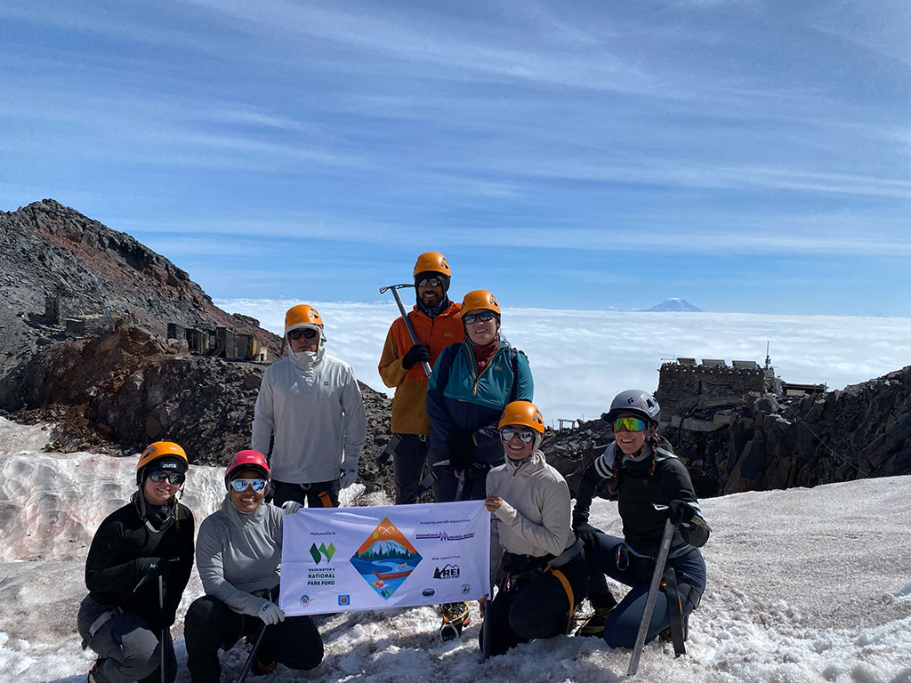 A group of Indigenous climbers hold a banner in front of a mountain vista