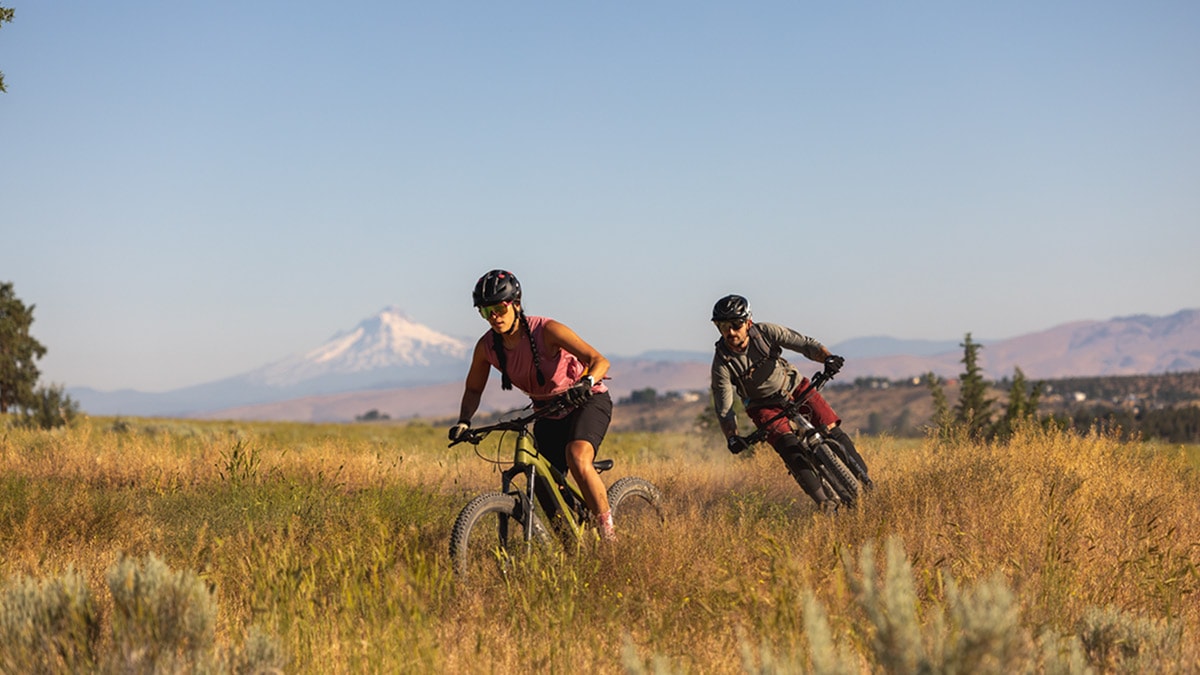 Two cyclists riding in a field against the backdrop of a mountain. 