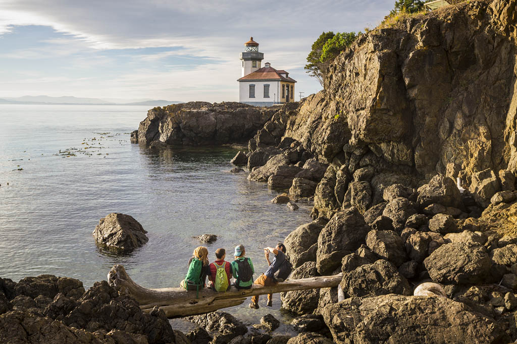 A group of people sit on a log overlooking a lighthouse.