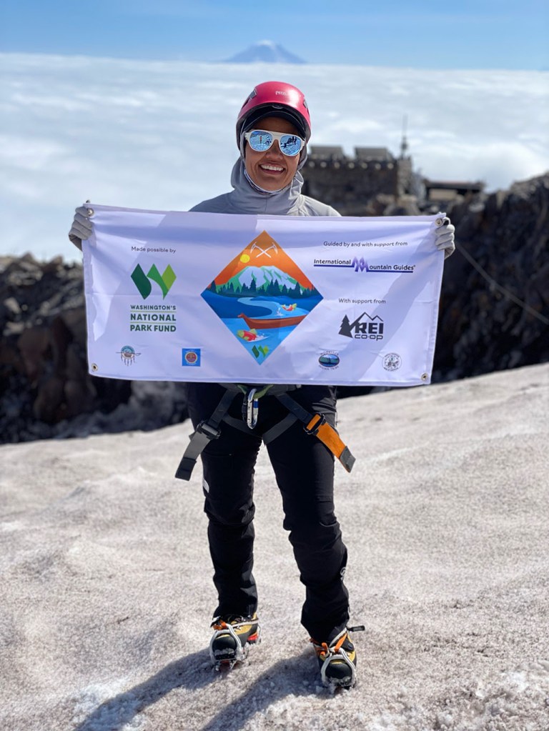 A portrait of Rachel Heaton, an Indigenous mountaineer, holding a banner while standing on a mountain