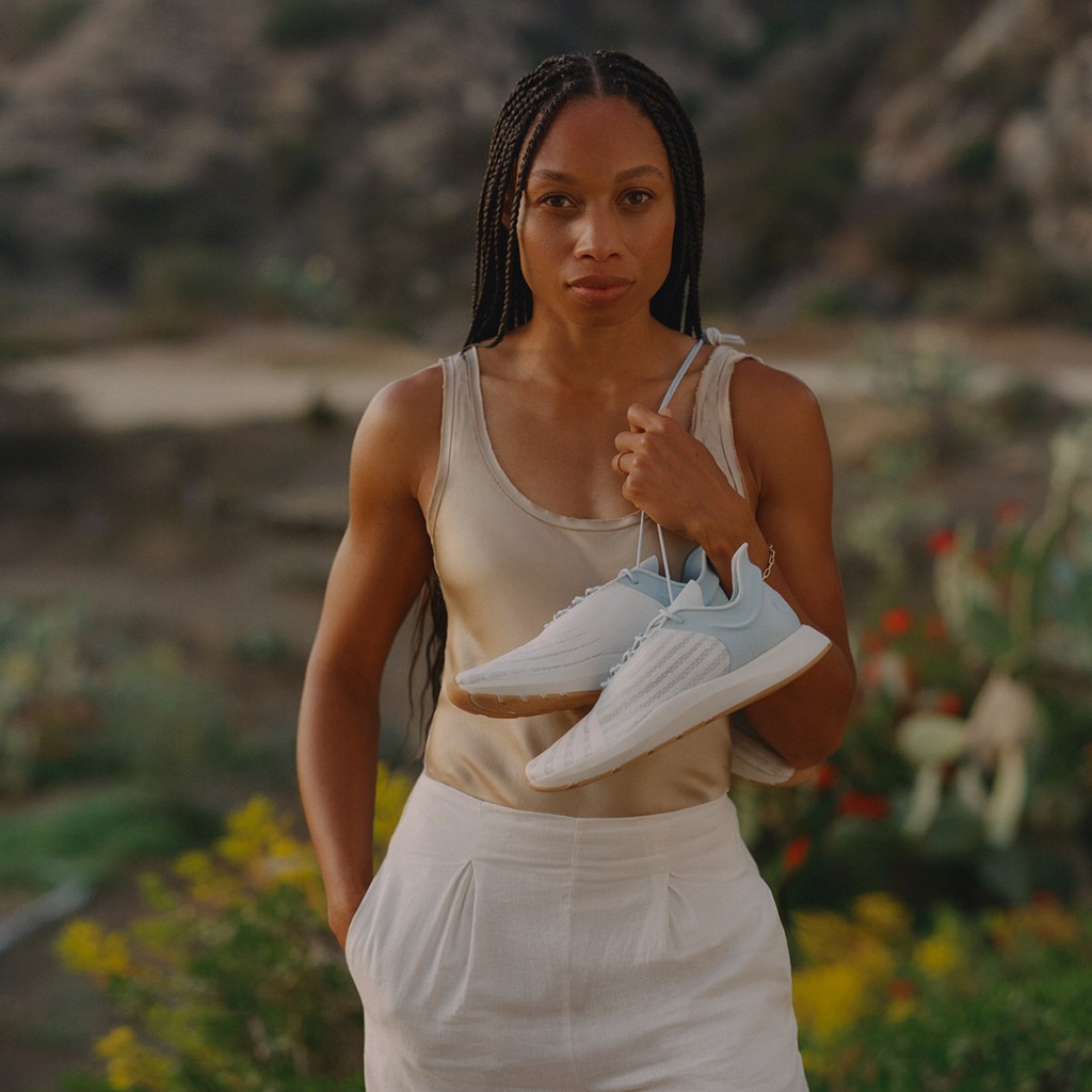 Saysh founder Allyson Felix standing in front of a natural backdrop holding a pair of sneakers