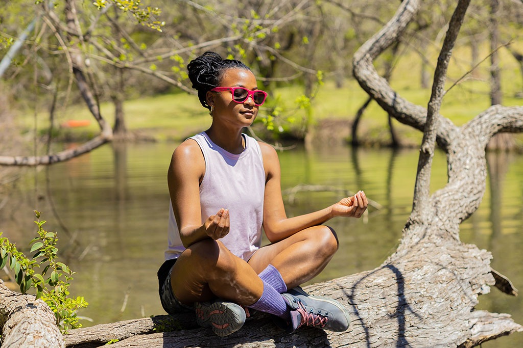 A Black hiker sits cross-legged on a log by a body of water.