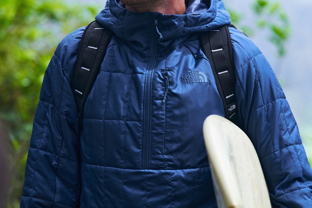 A person is wearing a Circaloft jacket and a backpack, carrying a surfboard.