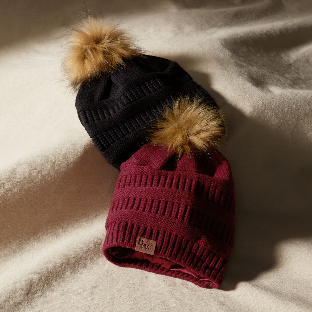 Two knit hats with fur pompoms lay on a cloth backdrop