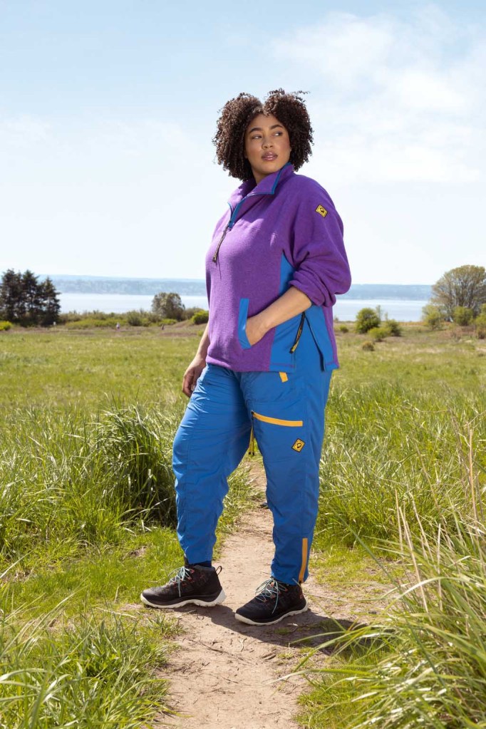 A person wearing a purple fleece top and bright blue pants with yellow zippers standing in a field