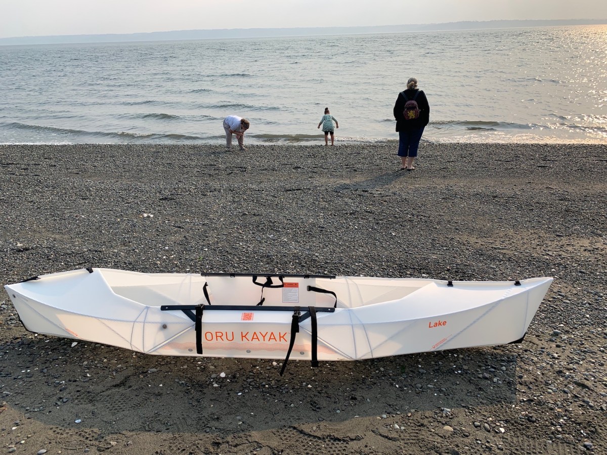 The author's empty Oru Kayak Lake Kayak resting on a pebble beach. A  family of three stands ahead of it with their backs to the camera, approaching the water. 