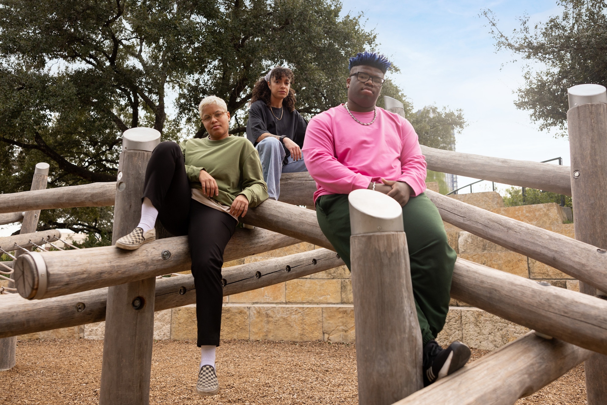 Three people dressed in the REI Co-op Active Pursuits collection, sitting together on a wooden structure in relaxed poses and looking at the camera.