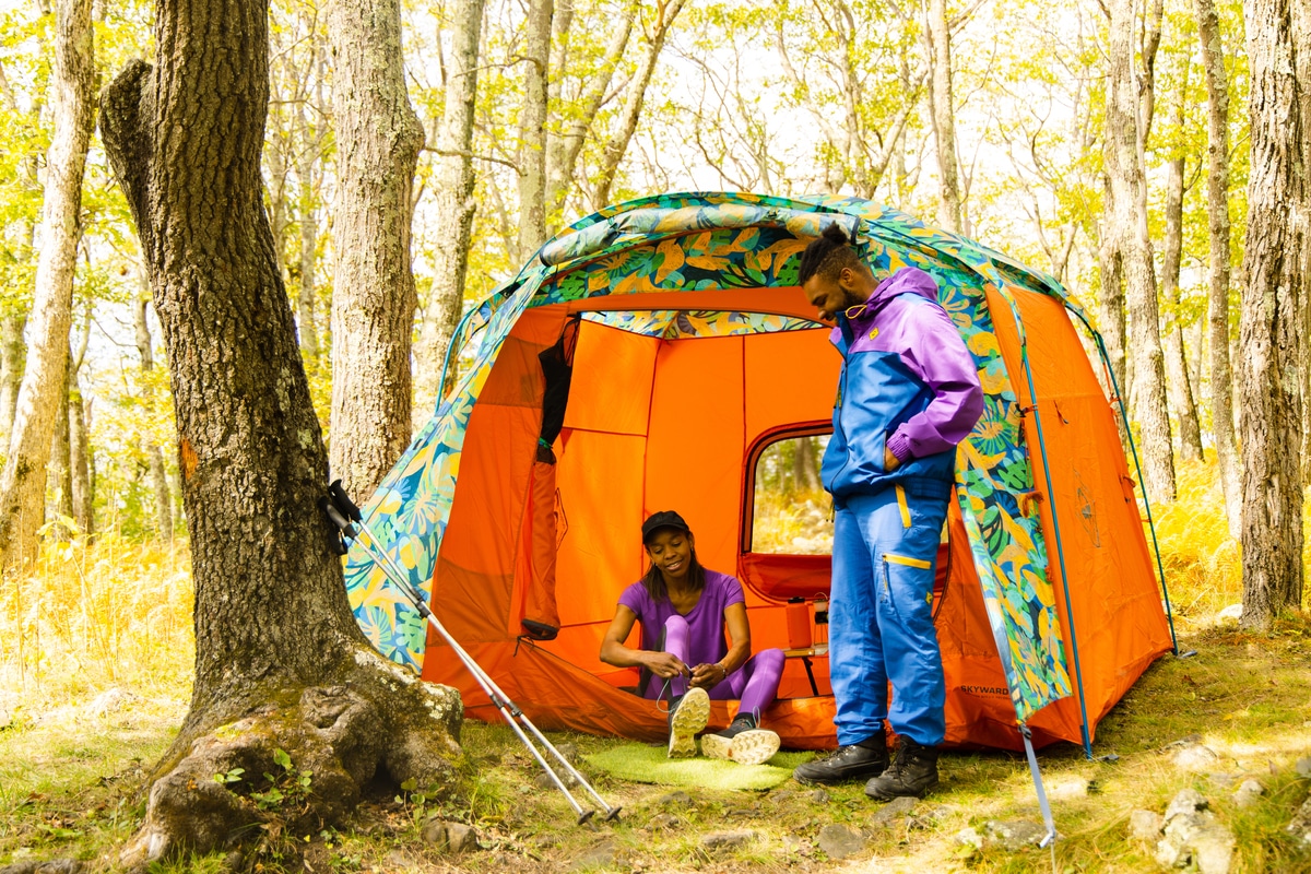 Two people wearing colorful Outdoor Afro + REI Co-op clothes stand in front of a brightly colored tent with a patterned fly designed by Chelsea Alexander