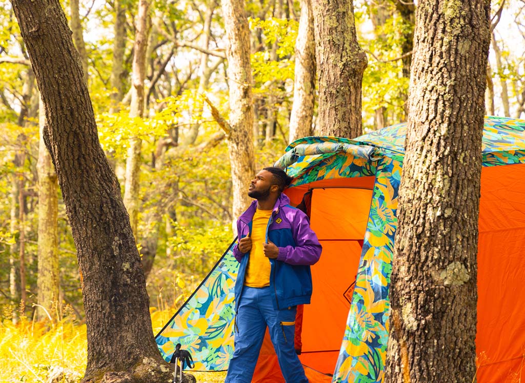 A Black camper wearing colorful outdoor clothes steps out of a brightly colored tent