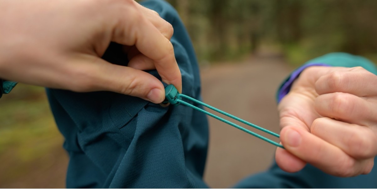 A model cinches the hood of the Flash Stretch rain jacket, in the Sea Forest/Crater Teal color way, which is a blue color, by holding the fastener in the back of the hood and pulling the drawstring.