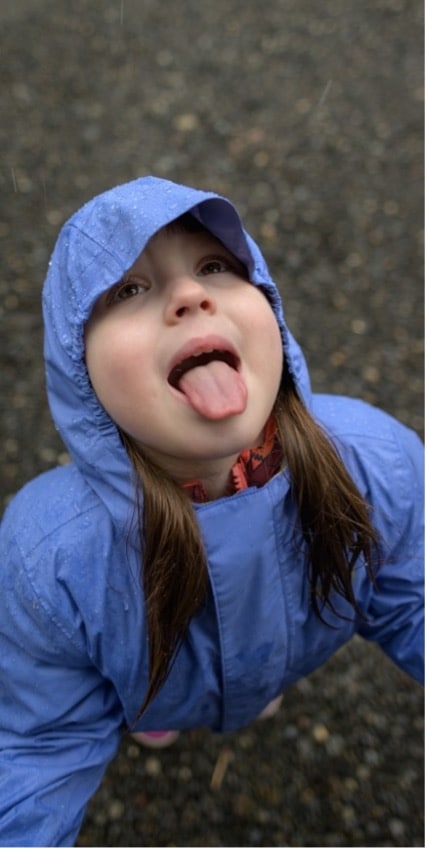 The author's daughter in the REI Co-op Rainier rain jacket for toddlers in Astral Purple. We see her from overhead looking up towards the rain and sticking her tongue out.