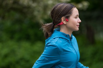 A woman in a blue rain jacket running in a pair of red Shokz OpenRun Headphones. We see her profile as she runs past, and there are green trees in the background.