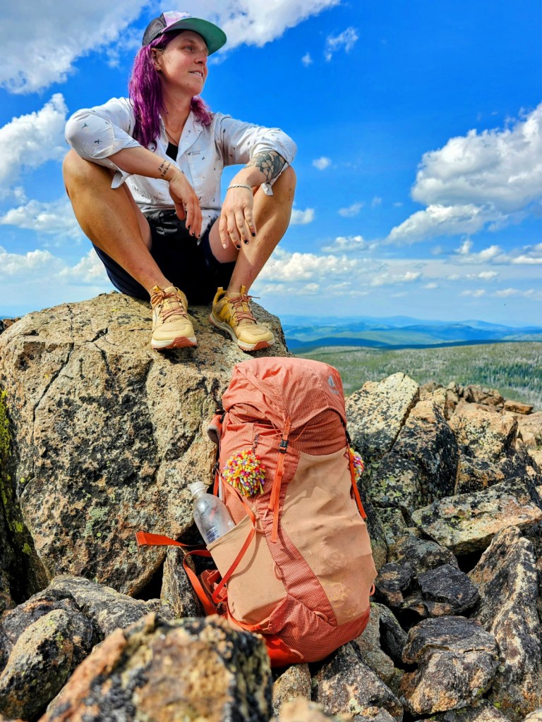 Lyla Harrod sits on a rock with an orange pack featuring a colorful puff ball.
