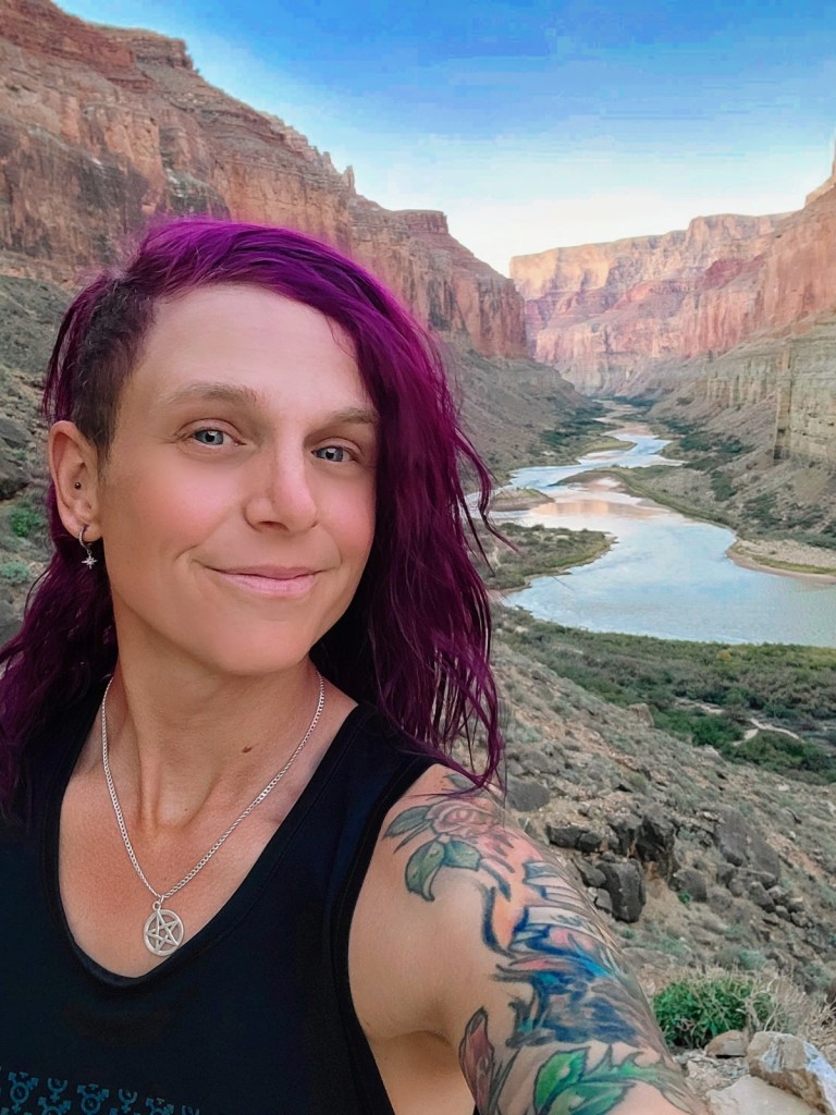 A person with shoulder-length dyed-purple hair looks into the camera for a selfie in front of a river that runs through the base of a canyon.
